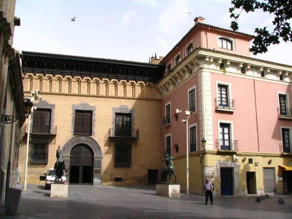 Historic European courtyard with yellow brick and pink buildings, equestrian statues, and people walking.