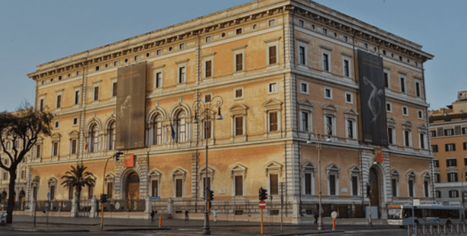 Large historic four-story building with arched windows, symmetrical architecture, palm trees, and banners.