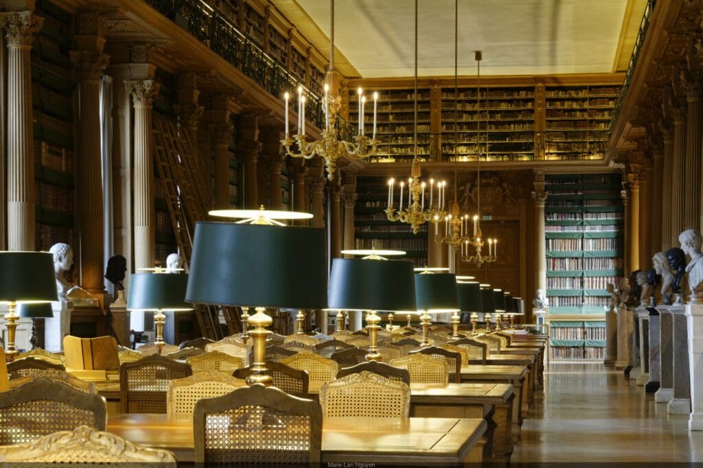Historic library interior with desks, green-shaded lamps, wooden chairs, chandeliers, and busts.