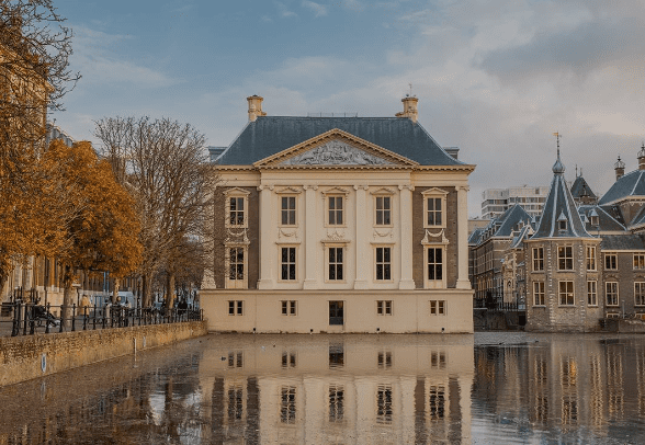 Historic neoclassical building with ornate detailing by calm water, surrounded by autumn trees.