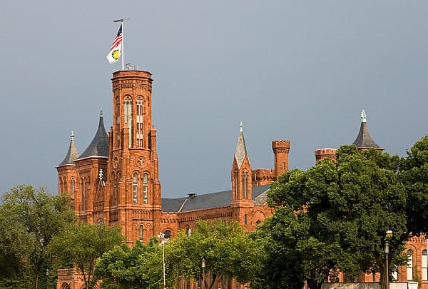 Historic red-brick building with towers and lush green trees. Central spire has two flags.