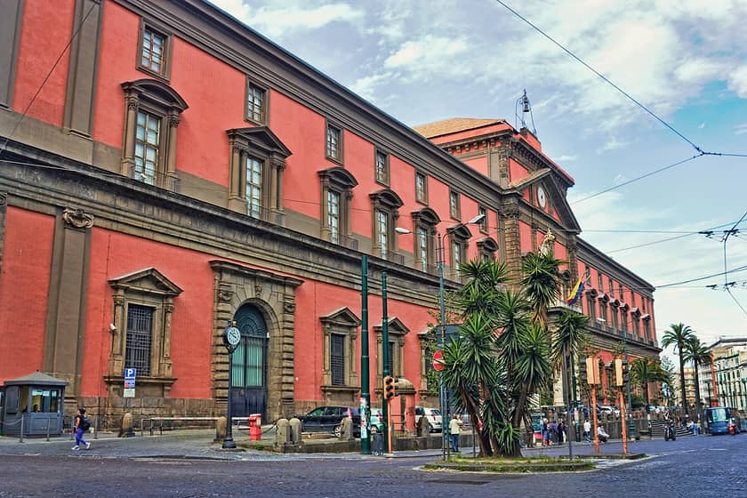 Historic red building with ornate details, palm trees, streetlamps, pedestrians, and partly cloudy sky.
