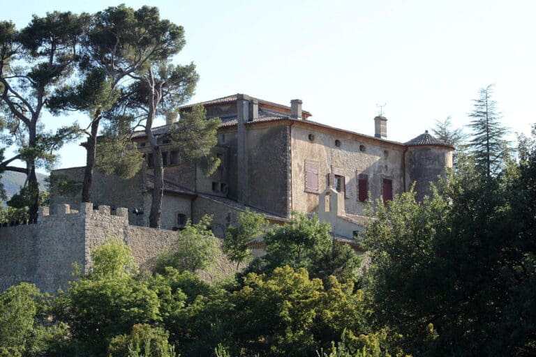 Historic stone building with red shutters, chimneys, and a stone wall, surrounded by tall pine trees.