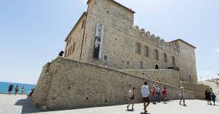 Historic stone building with arched windows and banner, visitors nearby, sea and blue sky background