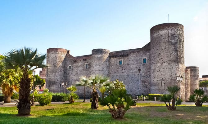 Historic stone castle with cylindrical towers, surrounded by palm trees and greenery, under clear sky.