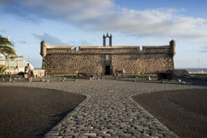 Historic stone fort with cylindrical watchtowers by a waterfront, cobblestone pathway, clear sky