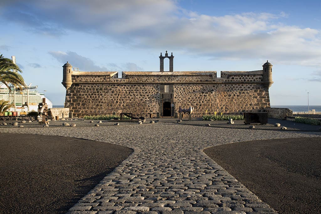 Historic stone fort with cylindrical watchtowers by a waterfront, cobblestone pathway, clear sky