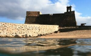 Historic stone fortress on sandy shore with cannon and bell tower, partially reflected in water.