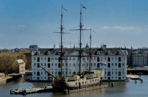 Historic tall ship moored at a pier, next to a multi-story white building with a gabled roof.