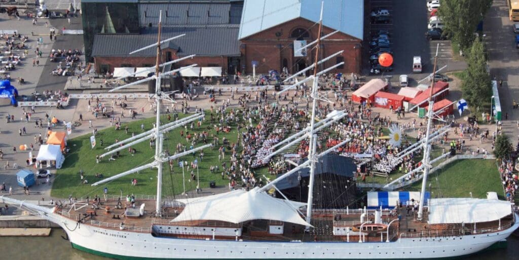 Aerial view of a historic tall ship docked, with people and stalls at a waterfront event area.