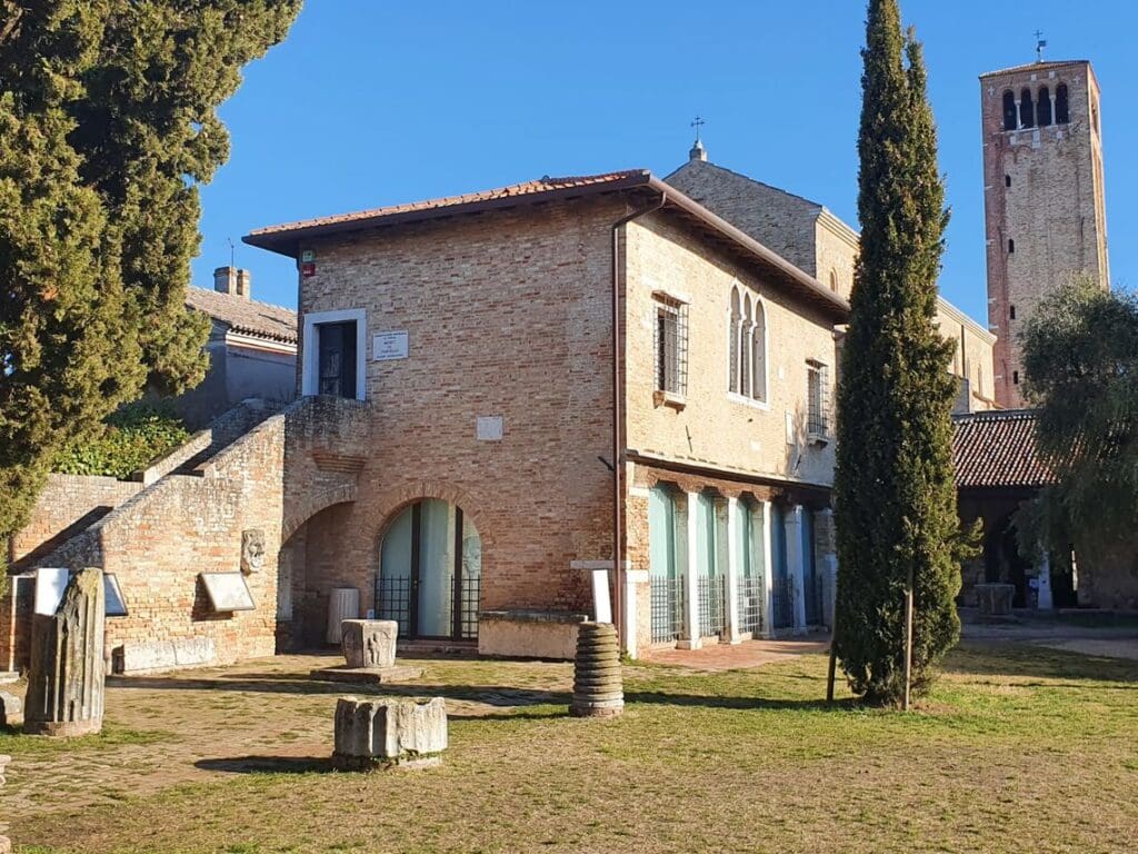 Historical brick building with arched doors, stone stairs, artifacts, and a tall brick tower in background