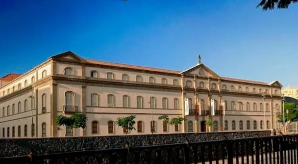 Large two-story historical building with arched windows, central pediment, trees, and a fence.