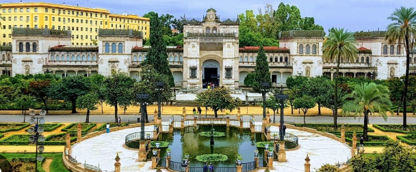 Historical building with ornate classical façade, lush garden, circular pond, framed by palm trees