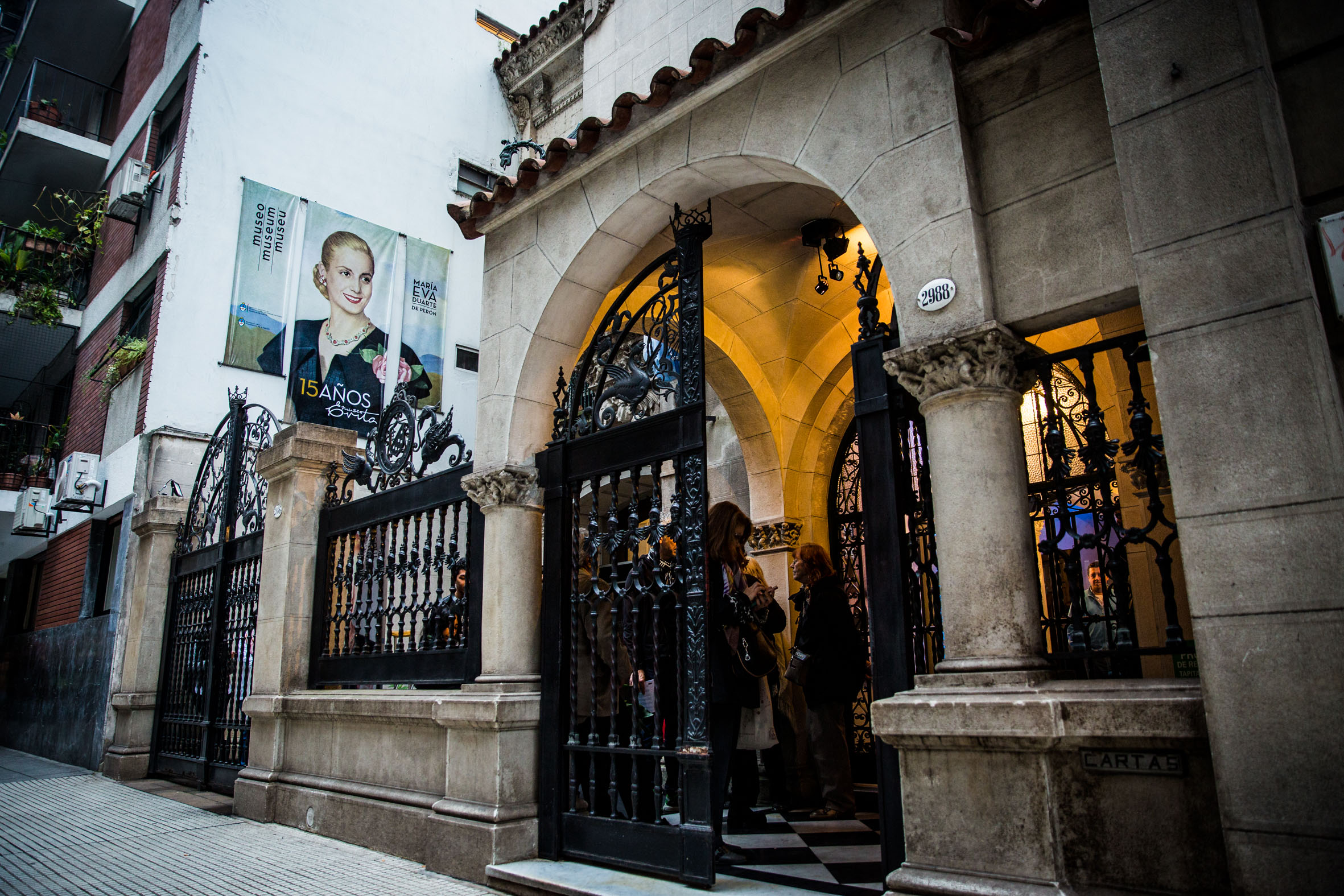 Historical building entrance with stone arch, black iron gate, museum poster, and people inside.