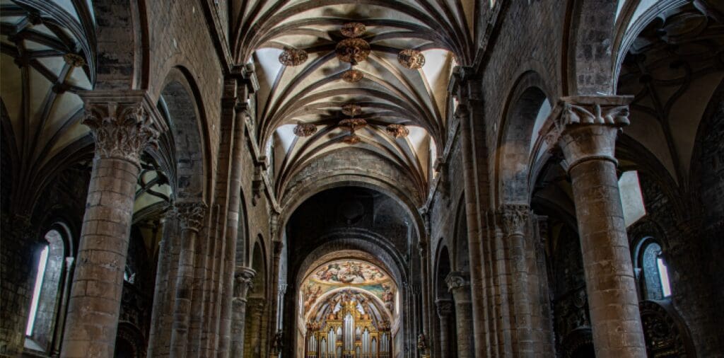 Interior of historical cathedral with stone columns, vaulted ceiling, high windows, and decorated altar.