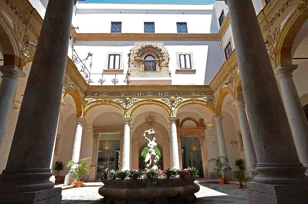 Historical courtyard with Italian-style arches, marble statue, plants, fountain, and intricate detailing.