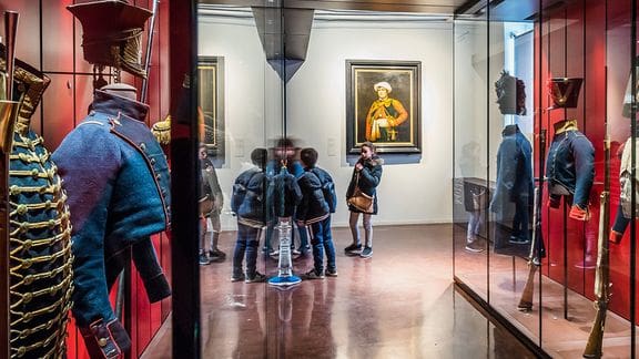 Historical military uniforms on display in glass cases at a museum exhibit with visitors observing.