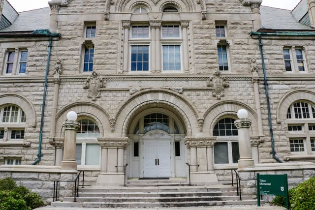 A historical stone building with arched entrance, columns, steps, and a green "Tilton Memorial" sign.