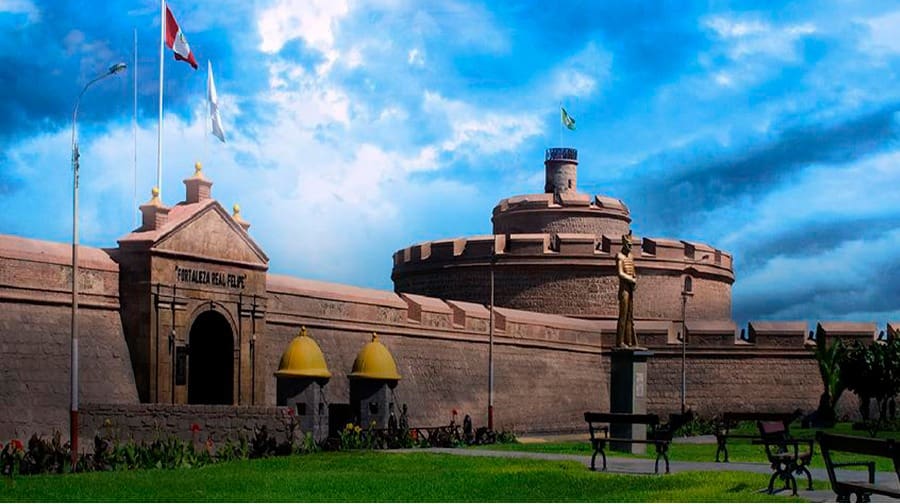 Historical stone fort with cylindrical tower, yellow-domed entrance, flags, statue, and landscaped park.