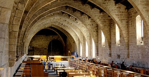 Spacious historical stone hall library with arched ceilings, wooden shelves, narrow windows, and people.