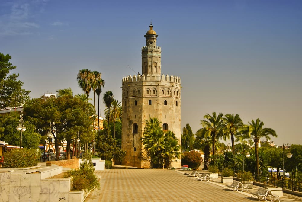 Historical stone tower with cylindrical shape, arched windows, surrounded by greenery and palm trees.