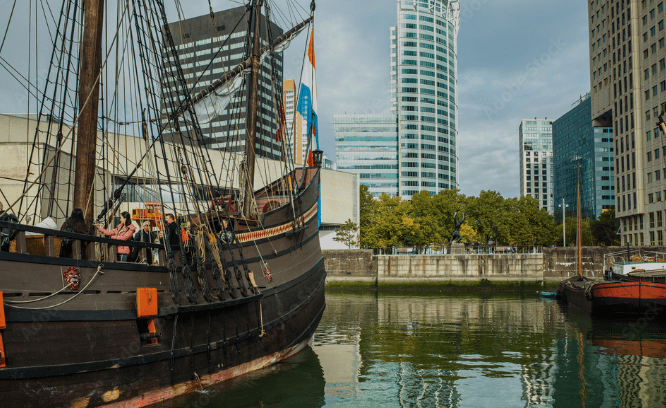 Historical wooden ship docked in a modern city harbor, surrounded by tall buildings and trees.