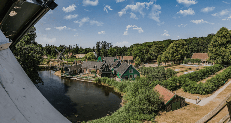 Idyllic rural scene with traditional green houses, water, lush trees, winding paths, and windmills.