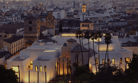 Illuminated complex with modern and historic buildings, tall palm trees, and urban cityscape at dusk