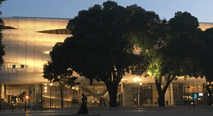 Illuminated, modern building partly obscured by trees with street lamps and pedestrian at twilight.