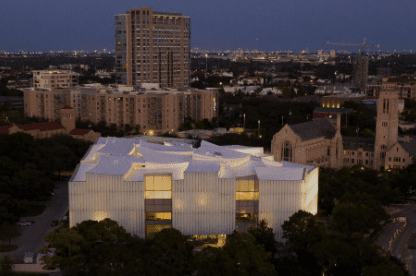 Illuminated modern building with wavy roof, large windows, surrounded by trees, homes, and a church.