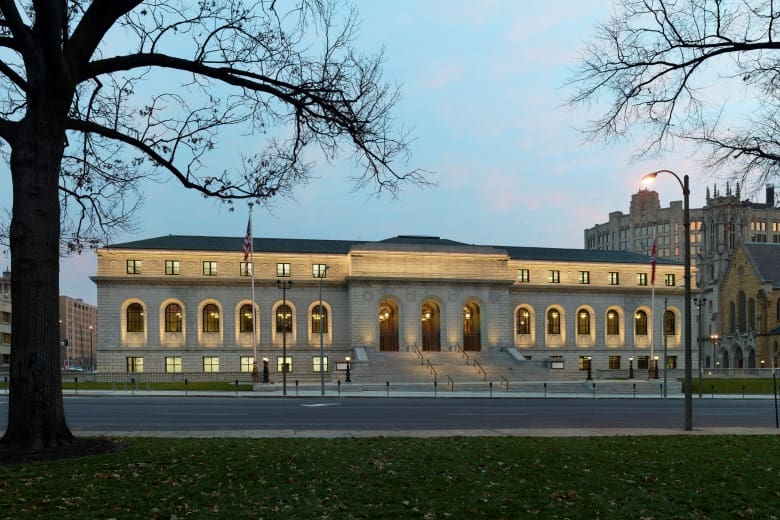 Illuminated neoclassical building with arched windows, stairs, and flagpoles, seen from a street.