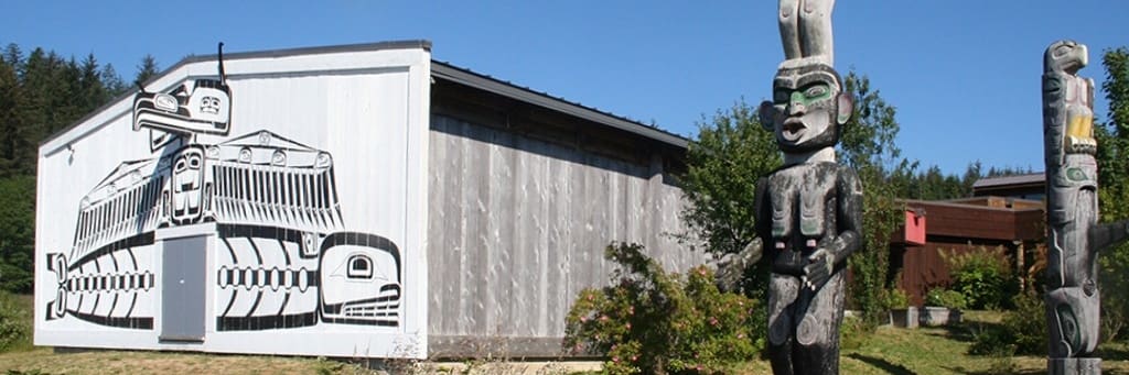Building with indigenous mural, two carved totem poles, garden area, and distant trees.