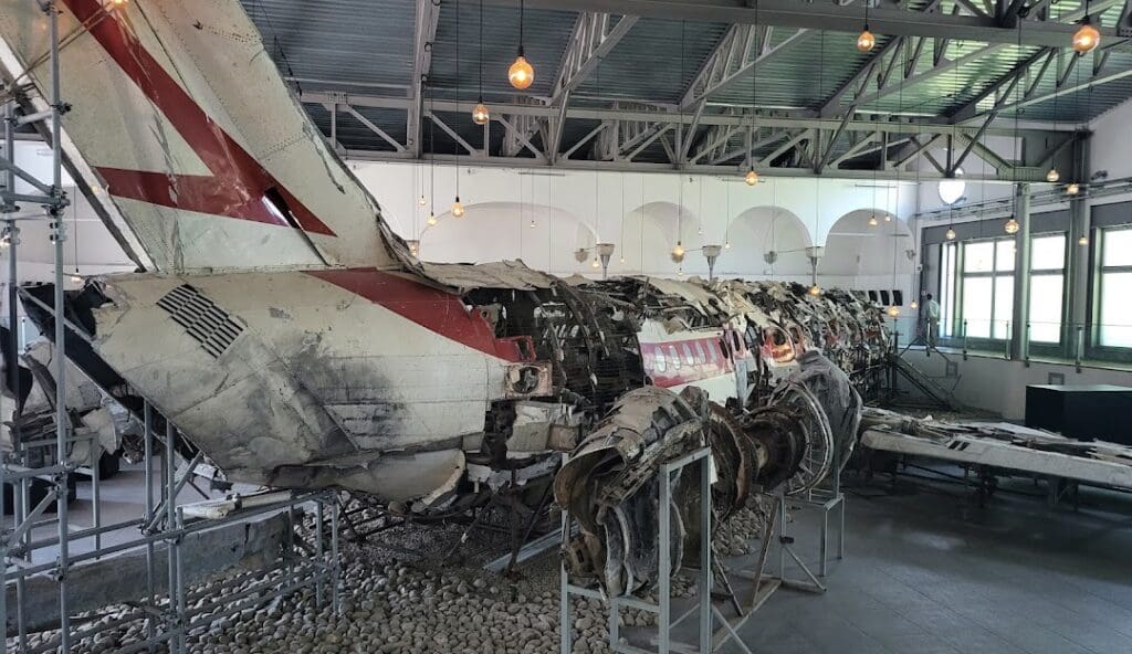 Indoor exhibit showcasing wreckage of an aircraft fuselage with support structures and natural light