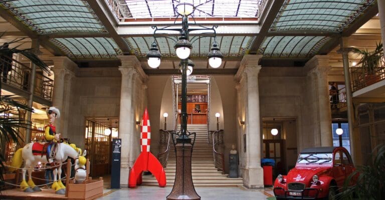 Indoor space with glass ceiling, decorative lighting, central staircase, statue, red car, and rocket model.
