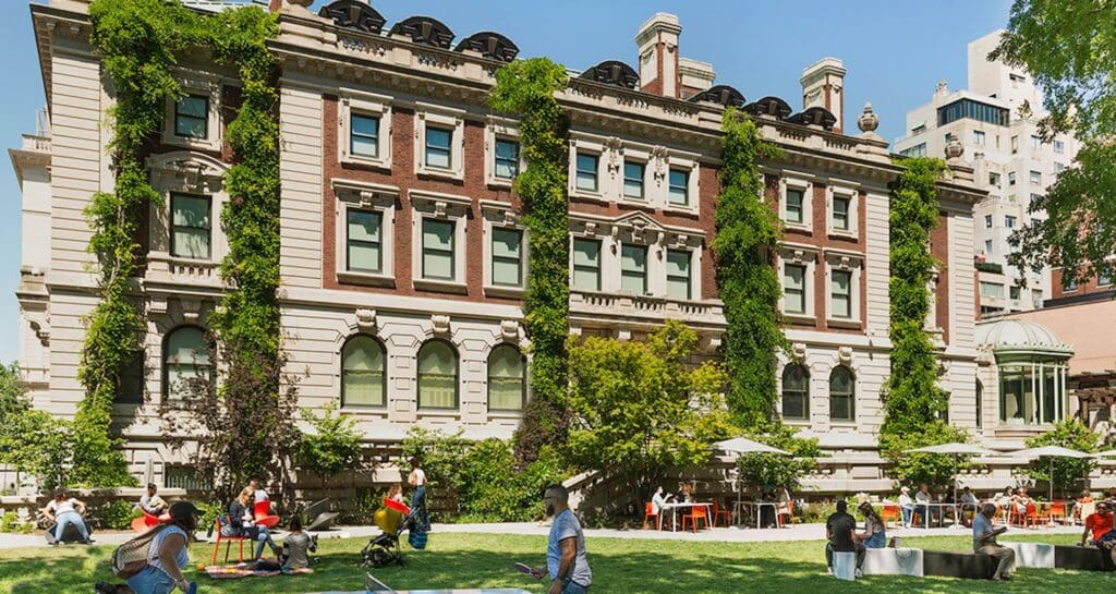 Ivy-covered multi-story building with classical architecture, surrounded by people on a green lawn.