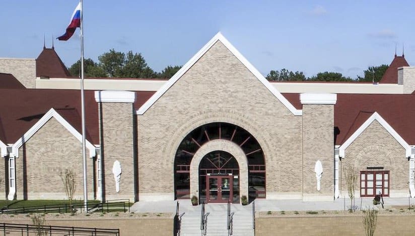 Large beige-brick building with red roof, arched main entrance, flagpole, stairs, and surrounding greenery.