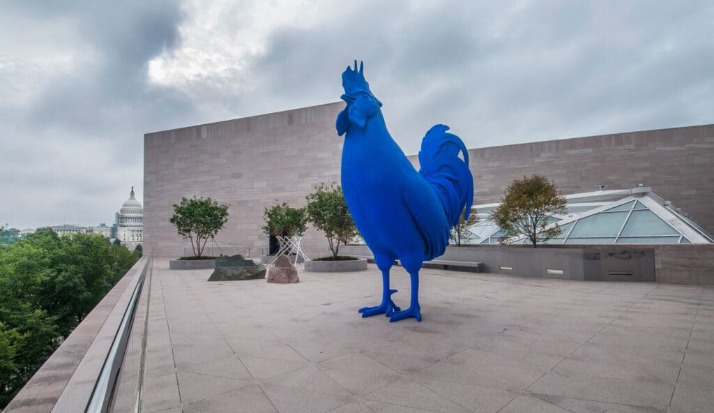 Large blue rooster sculpture on a rooftop terrace with distant dome-shaped building under cloudy sky.