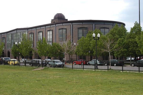 Large curved brick building with windows, trees, fence, parked cars, and a grassy foreground.