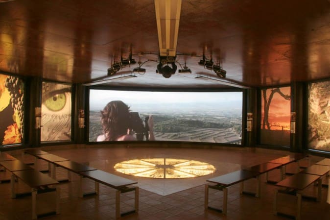 Large curved screen showing person using camera; benches, compass design floor, and art in the room.