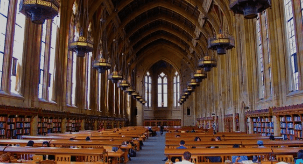 Large, well-lit library with high ceilings, arched windows, wooden tables, bookshelves, and chandeliers.