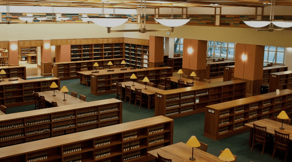 Large library interior with wooden bookshelves, desks, reading lamps, and rows of seating areas.