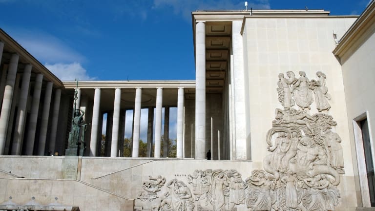 Large structure with tall white columns, detailed stone carvings, and a statue against clear sky.