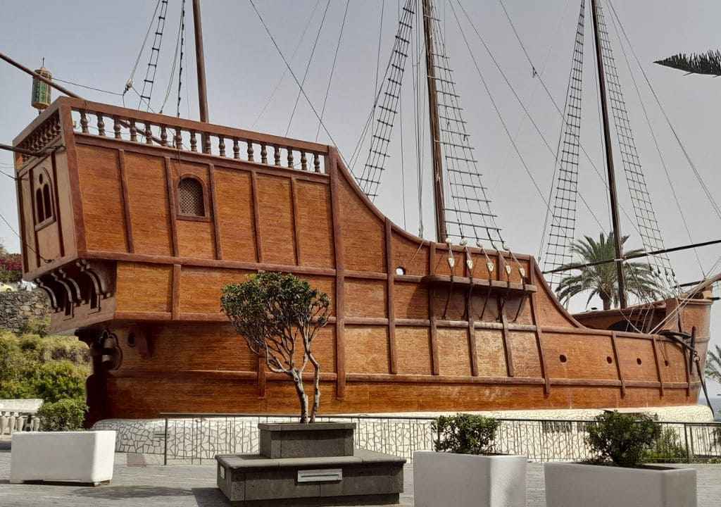 Large wooden ship with masts and rigging on outdoor display, surrounded by plants and benches.
