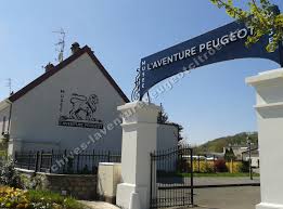 Entrance of L'Aventure Peugeot museum with arch, logo, fenced area, and garden in the background.