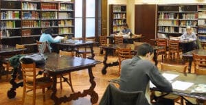 Library interior with people seated at wooden tables, reading and studying, shelves of books around.