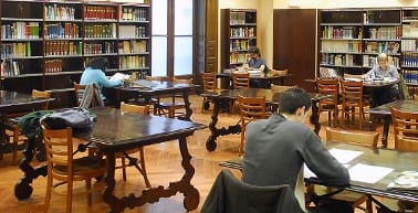 Library interior with people seated at wooden tables, reading and studying, shelves of books around.