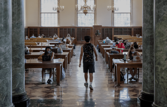 Reading room in a library with students studying at wooden tables, well-lit by windows and chandeliers.