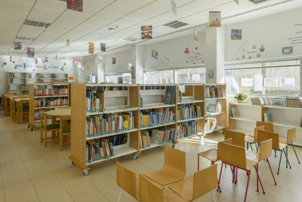 Well-lit library room with bookshelves, round tables, chairs, and large windows for natural light.