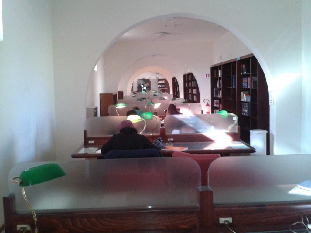 Library study area with wooden desks, glass dividers, green lamps, archways, and bookshelves.