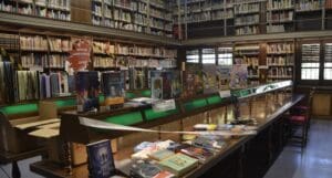 Library with a long wooden table displaying books under green lamps, surrounded by shelves.